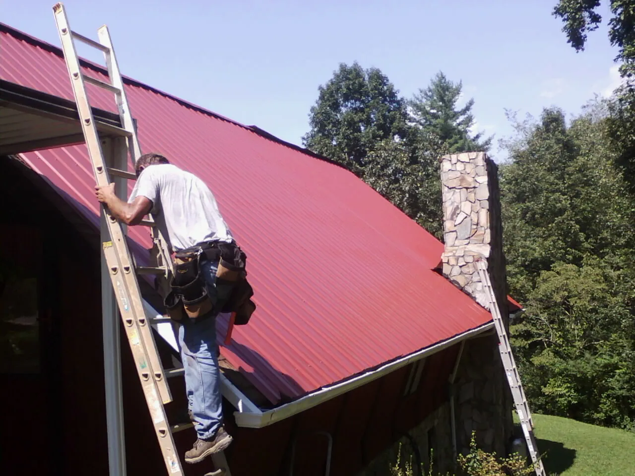 Roofer on a ladder beside a red metal roof and stone chimney — Soffit & Fascia Repair by Roofing Brothers in New Suffolk